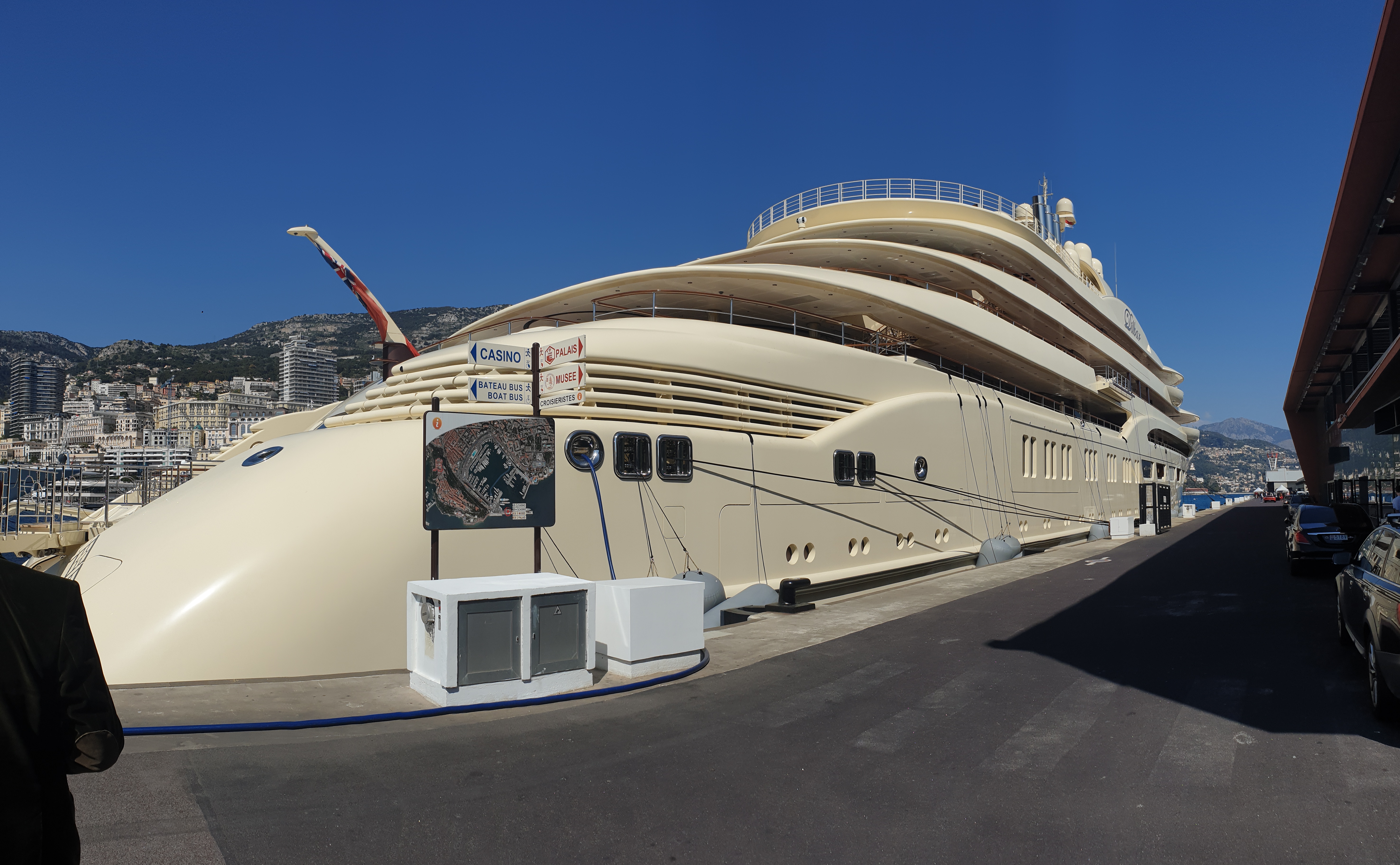 Large luxury yacht docked in a marina with cityscape and mountains in the background.