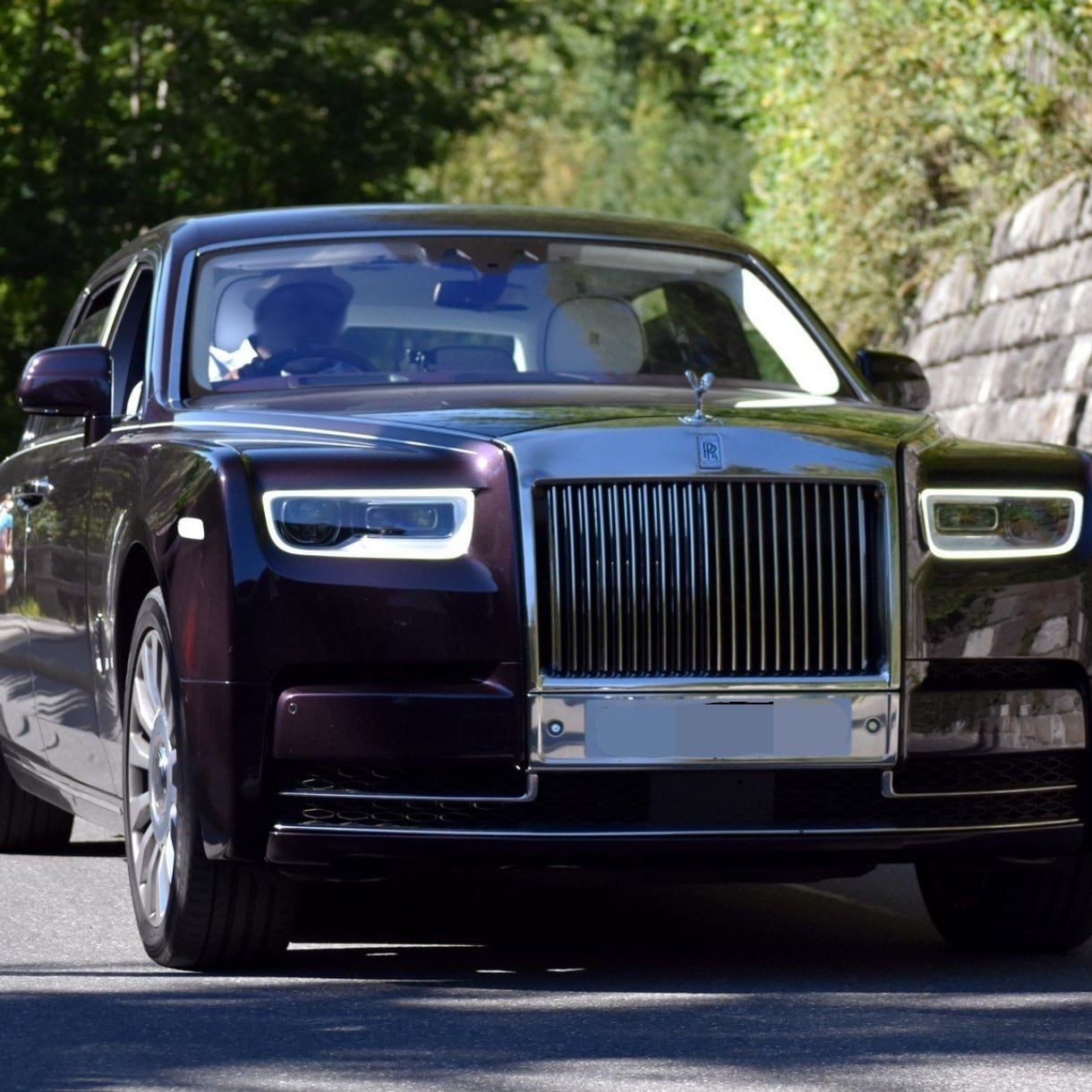 Front view of a black luxury car driving on a sunny road with trees in the background.